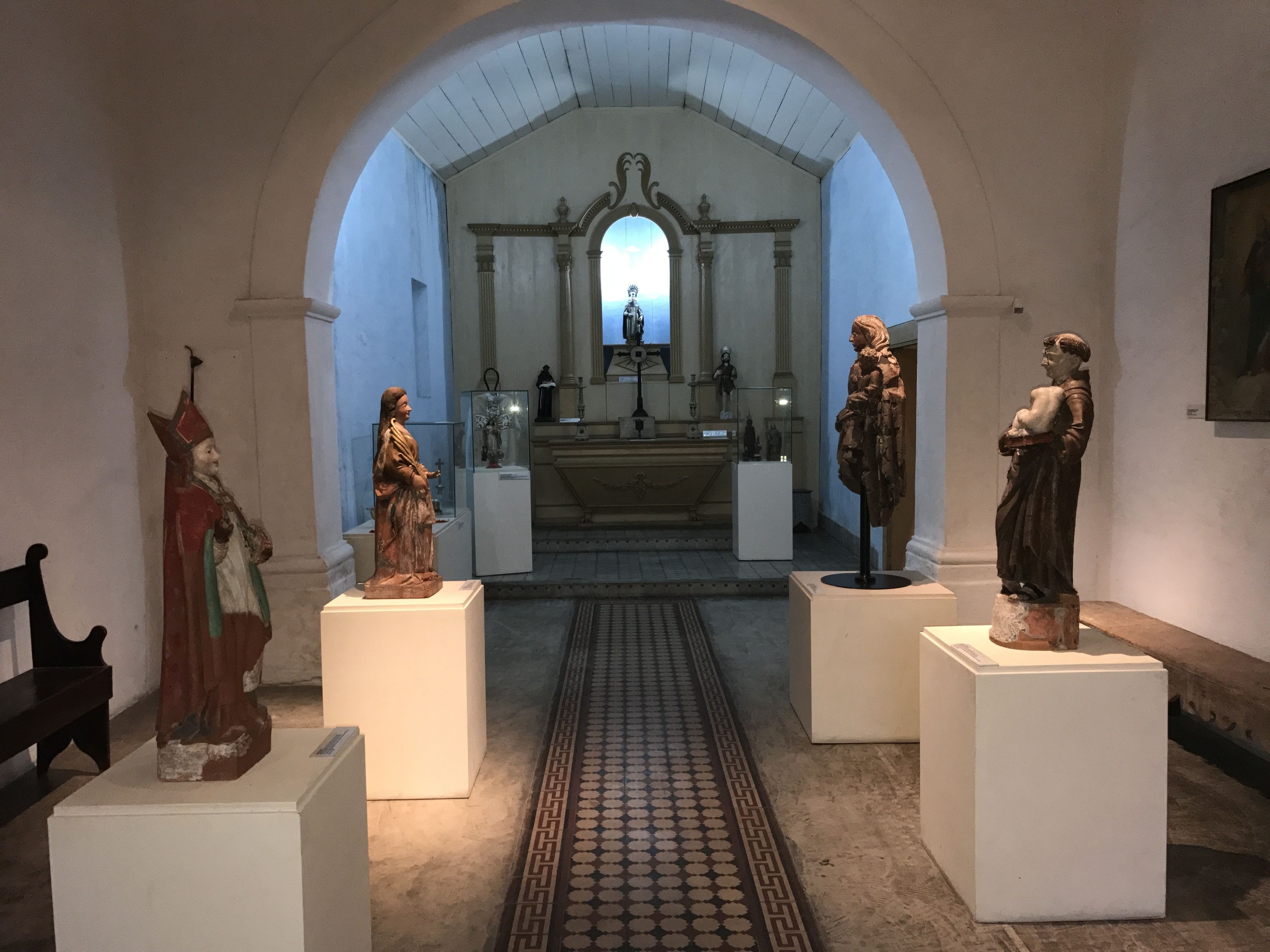 A museum interior with statues on pedestals, a central altar, and a patterned floor.