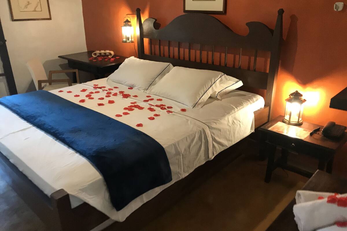 A neatly made bed with white linens and a blue blanket, a bedside table with a lamp, and a chair in a room with orange walls and framed pictures.