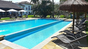 A swimming pool with lounge chairs and umbrellas, surrounded by palm trees and a building with a thatched roof. - Porto Grande Hotel & Convention (São Sebastião)