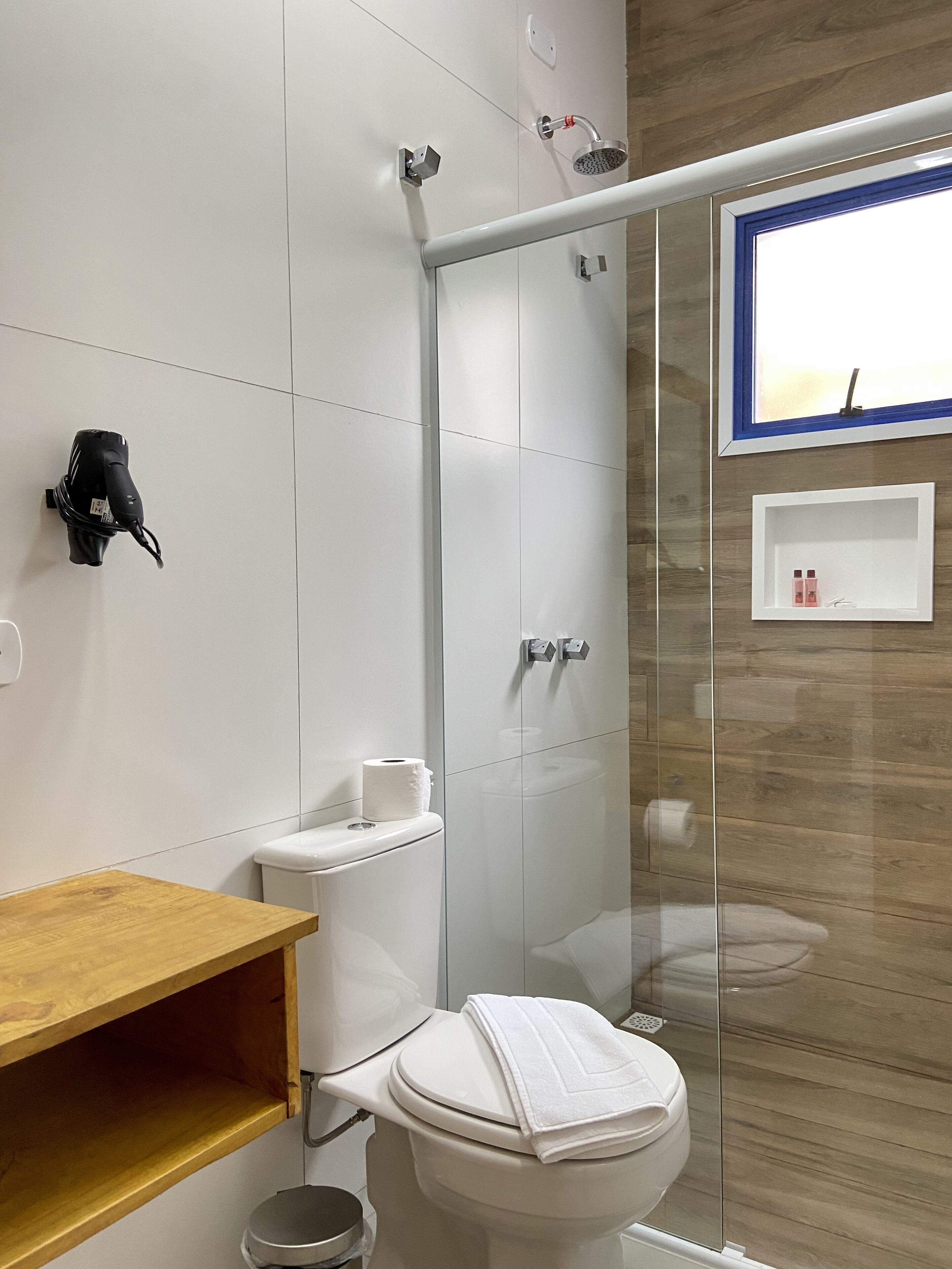Un baño moderno con cabina de ducha de vidrio, inodoro con una toalla encima, estante de madera y un pequeño estante blanco con artículos de tocador.