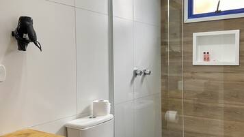 Un baño moderno con cabina de ducha de vidrio, inodoro con una toalla encima, estante de madera y un pequeño estante blanco con artículos de tocador.