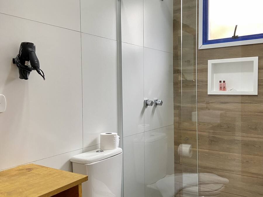 Un baño moderno con cabina de ducha de vidrio, inodoro con una toalla encima, estante de madera y un pequeño estante blanco con artículos de tocador.