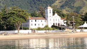A coastal building with a bell tower, surrounded by greenery and hills. - Porto Grande Hotel & Convention (São Sebastião)