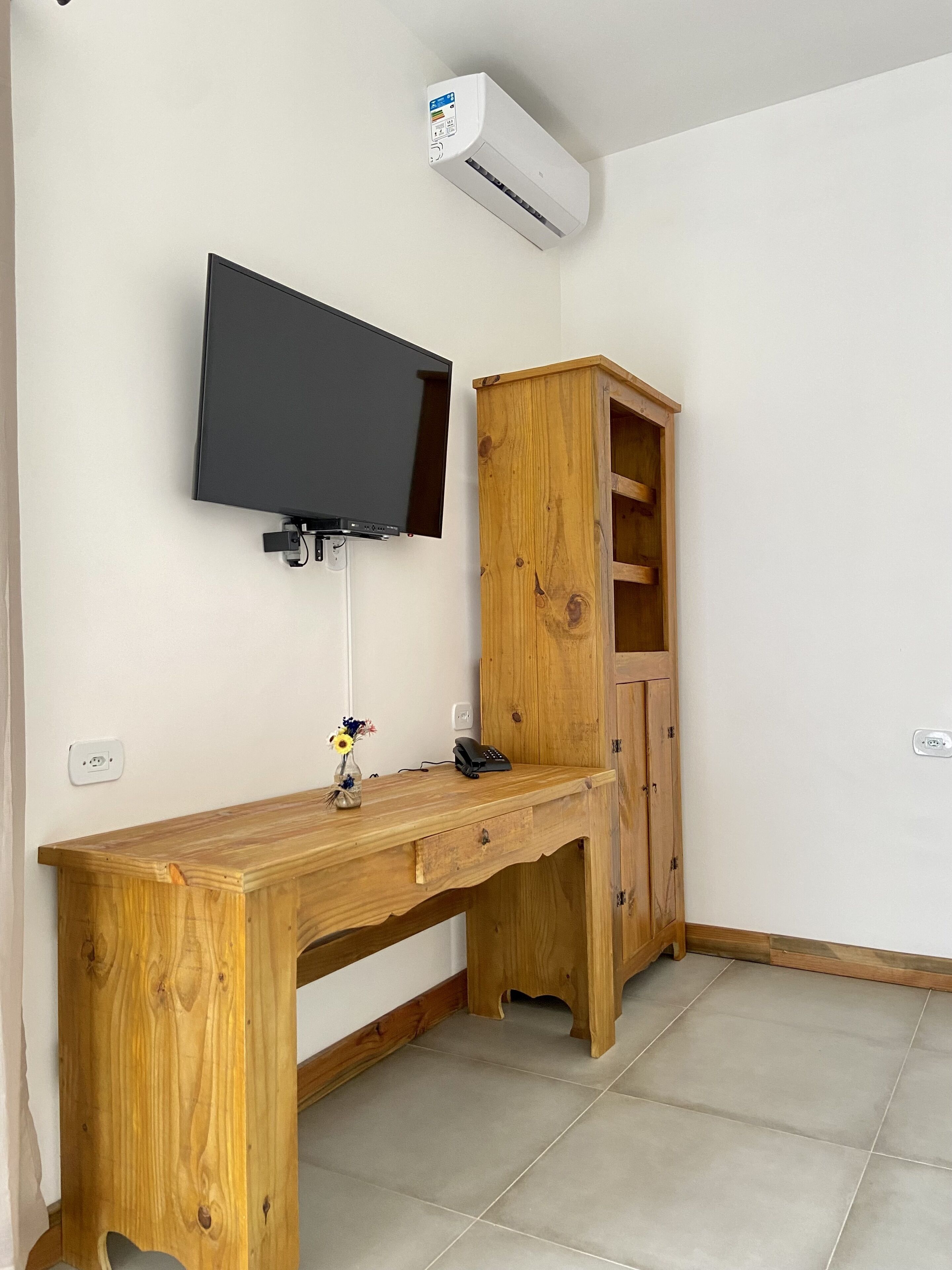 Una habitación con escritorio de madera, televisor de pantalla plana montado, unidad de aire acondicionado y mueble de madera.