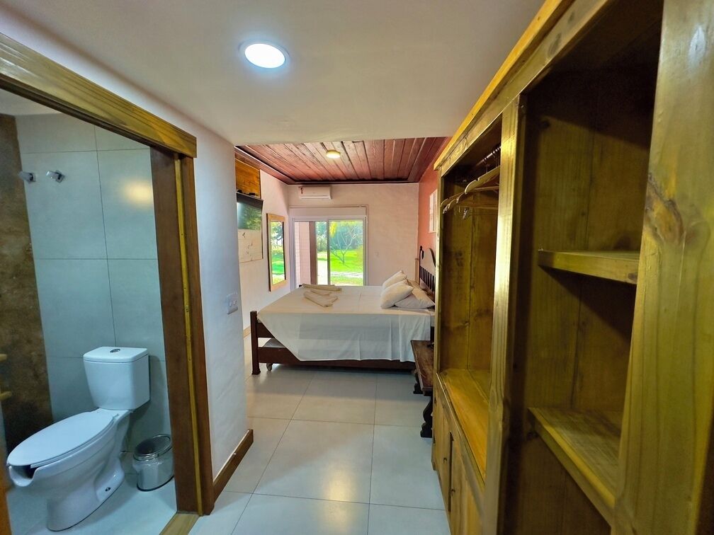 Hotelkamer met houten vloer, een bed met witte lakens, een wit toilet en een raam met uitzicht op groen.