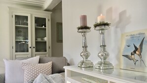 Un soggiorno con un divano bianco, cuscini decorati, un tavolino da caffè in vetro e due candele accese su supporti argentati. Sul tavolo è presente un libro intitolato "Die Vögel Europas".