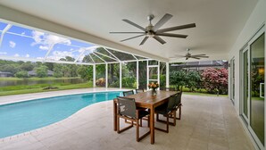 A covered patio with a pool, a wooden table with chairs, and ceiling fans. - Rovigo Vacation Rental: Spacious Retreat with Panoramic Lake Views in Kings Lake. 4-BR, 4-Bathroom Home with Private Pool, just 3.4 Miles from the Vibrant Fifth Avenue South and Naples Attractions (Naples)