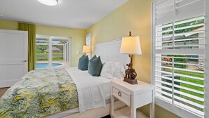 A bedroom with a bed, nightstand, lamp, and a view of a pool and garden through the window. - Rovigo Vacation Rental: Spacious Retreat with Panoramic Lake Views in Kings Lake. 4-BR, 4-Bathroom Home with Private Pool, just 3.4 Miles from the Vibrant Fifth Avenue South and Naples Attractions (Naples)