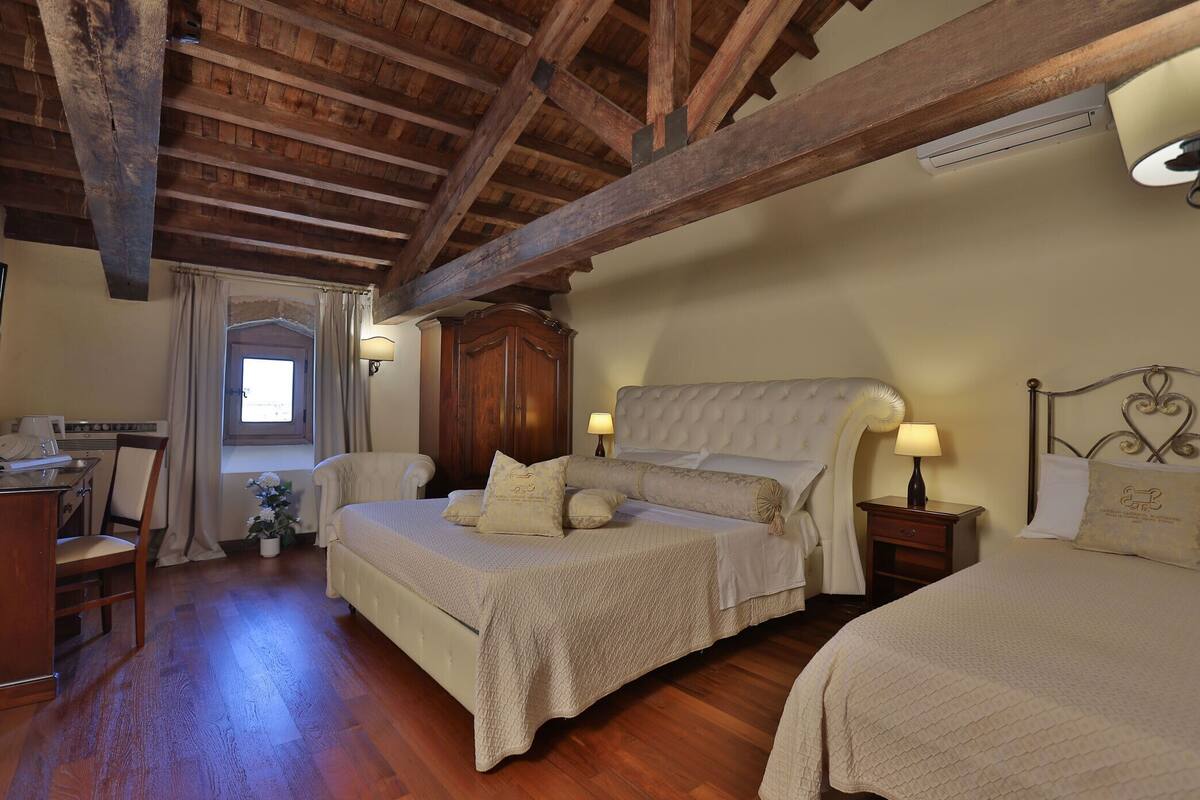 Une chambre avec un plafond en bois, un lit avec une tête de lit, des tables de chevet avec des lampes, un bureau avec une chaise et une fenêtre avec des rideaux.