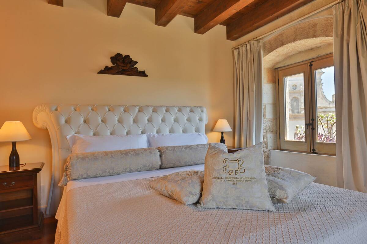 Une chambre à coucher avec une tête de lit capitonnée, un lit recouvert de draps blancs, deux lampes de chevet et une fenêtre avec des rideaux.