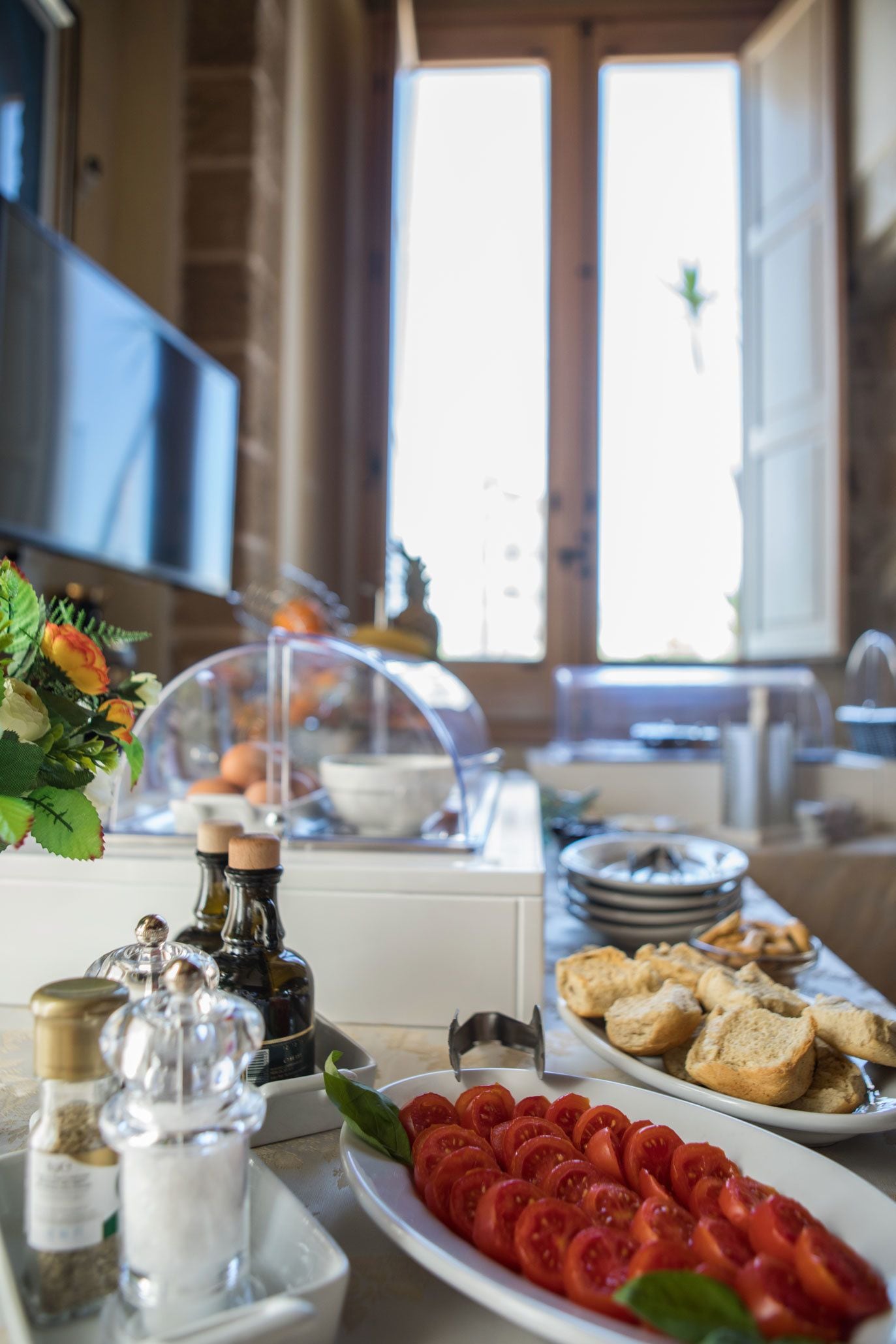 Un bufé de desayuno con diversos alimentos, incluyendo tomates en rodajas y pan, en una habitación bien iluminada con un amplio ventanal.