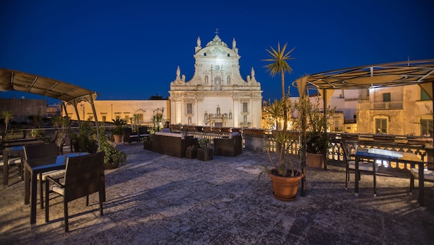 An outdoor patio area with seating and potted plants, a white building with ornate architecture in the background, and a palm tree to the right. - Castello Castriota Scanderbeg Suites De Charme (Galatina)