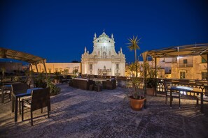 An outdoor patio area with seating and potted plants, a white building with ornate architecture in the background, and a palm tree to the right. - Castello Castriota Scanderbeg Suites De Charme (Galatina)