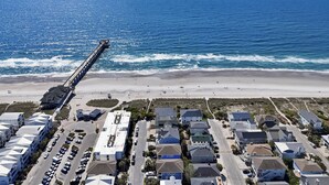An aerial view of a coastal area with a pier extending into the ocean, a beach, and a residential neighborhood with houses and parked cars.