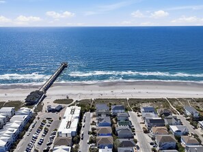 An aerial view of a coastal area with a pier extending into the ocean, a beach, and a residential neighborhood with houses and parked cars.