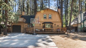 A two-story house with a basketball hoop in front, surrounded by tall trees.