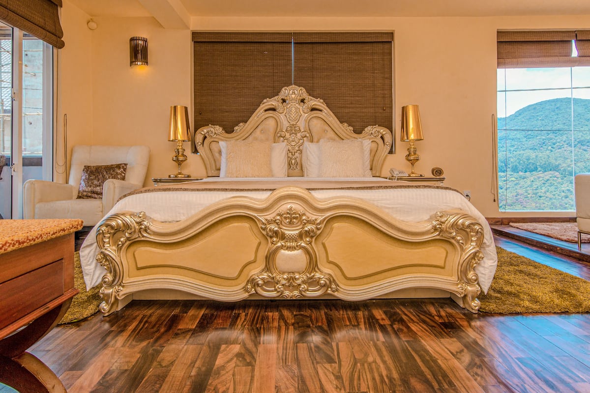 A large bed with ornate headboard and footboard, two bedside lamps, and a view of a mountainous landscape through a large window.