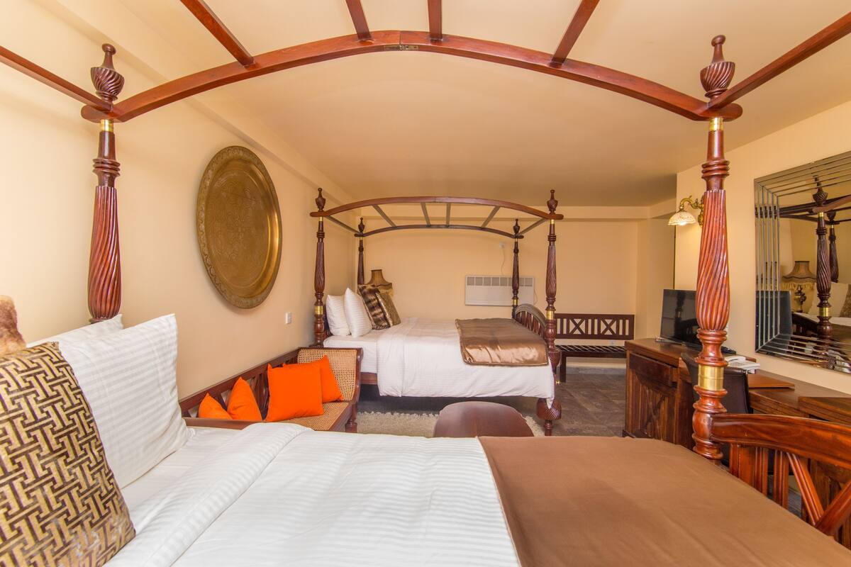 A four-poster bed with a wooden canopy, two single beds, a sofa with orange cushions, a desk, and a mirror.