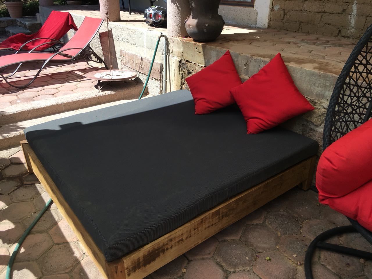 A wooden bed with a black mattress and red pillows, positioned on a stone patio with lounge chairs and a potted plant in the background.