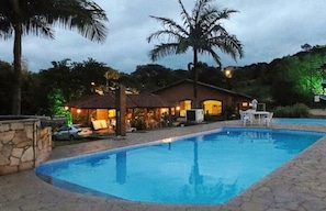 A swimming pool with a stone border, a house with a tiled roof, and palm trees. - Hotel Fazenda Reviver (Araçoiaba da Serra)