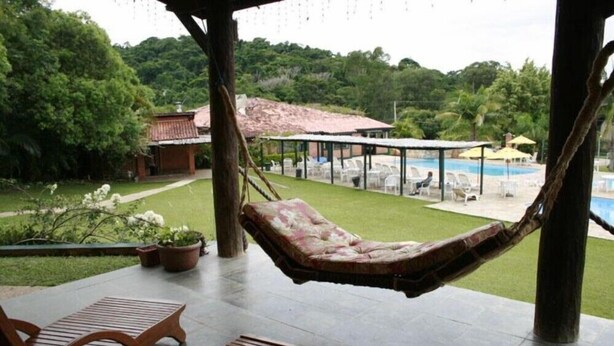 A hammock chair on a covered patio with a pool and lounge chairs in the background. - Hotel Fazenda Reviver (Araçoiaba da Serra)