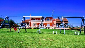 A grassy area with a hammock, swing set, and a red building in the background. - Riposo Studios & Apartments (Corfu)