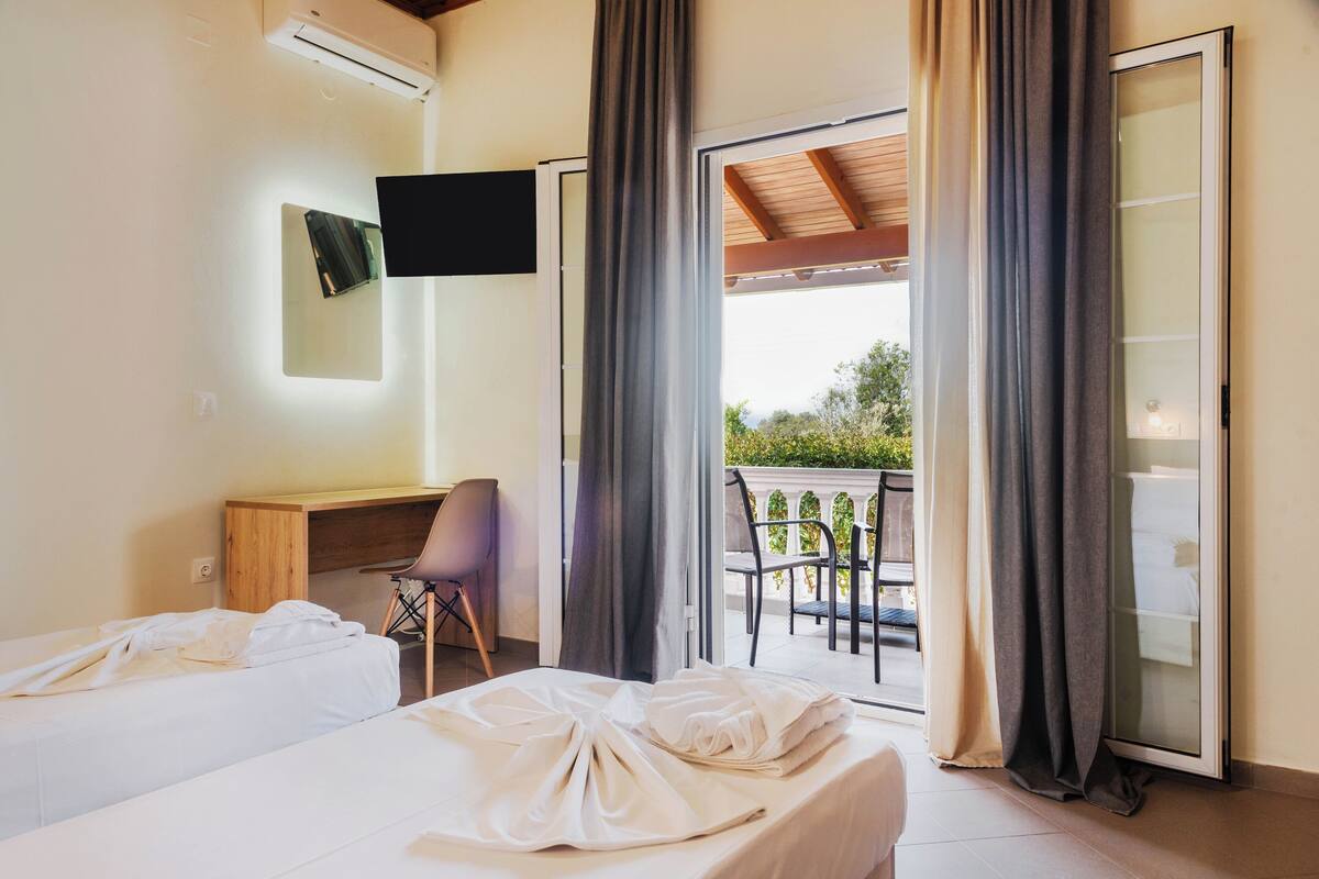 Une chambre d’hôtel avec deux lits, un bureau, une chaise, une télévision et un balcon avec des rideaux.