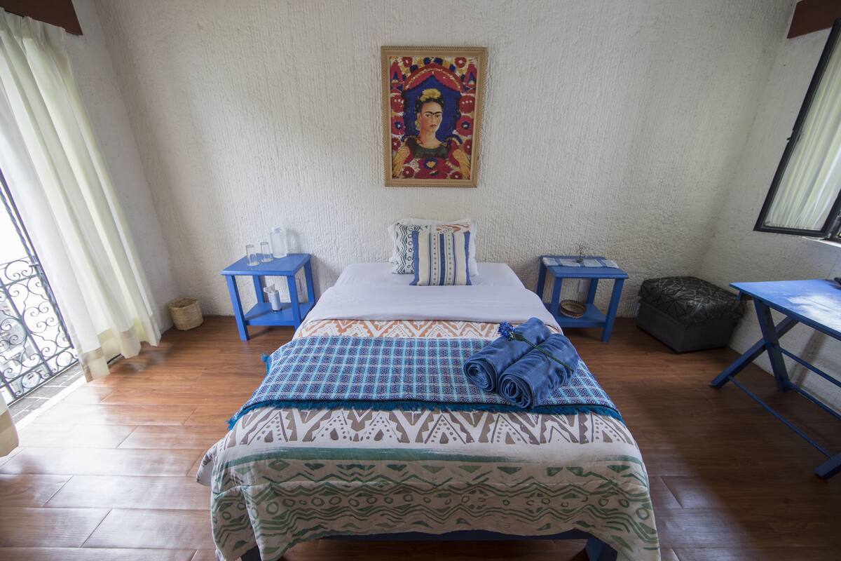 Un dormitorio con cama, dos mesitas de noche, una ventana con cortinas y un cuadro en la pared.