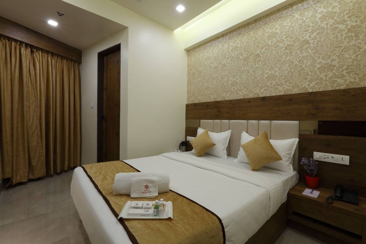 A hotel room with a neatly made bed, a bedside table with a phone and a red pot, and a wall with a patterned design.