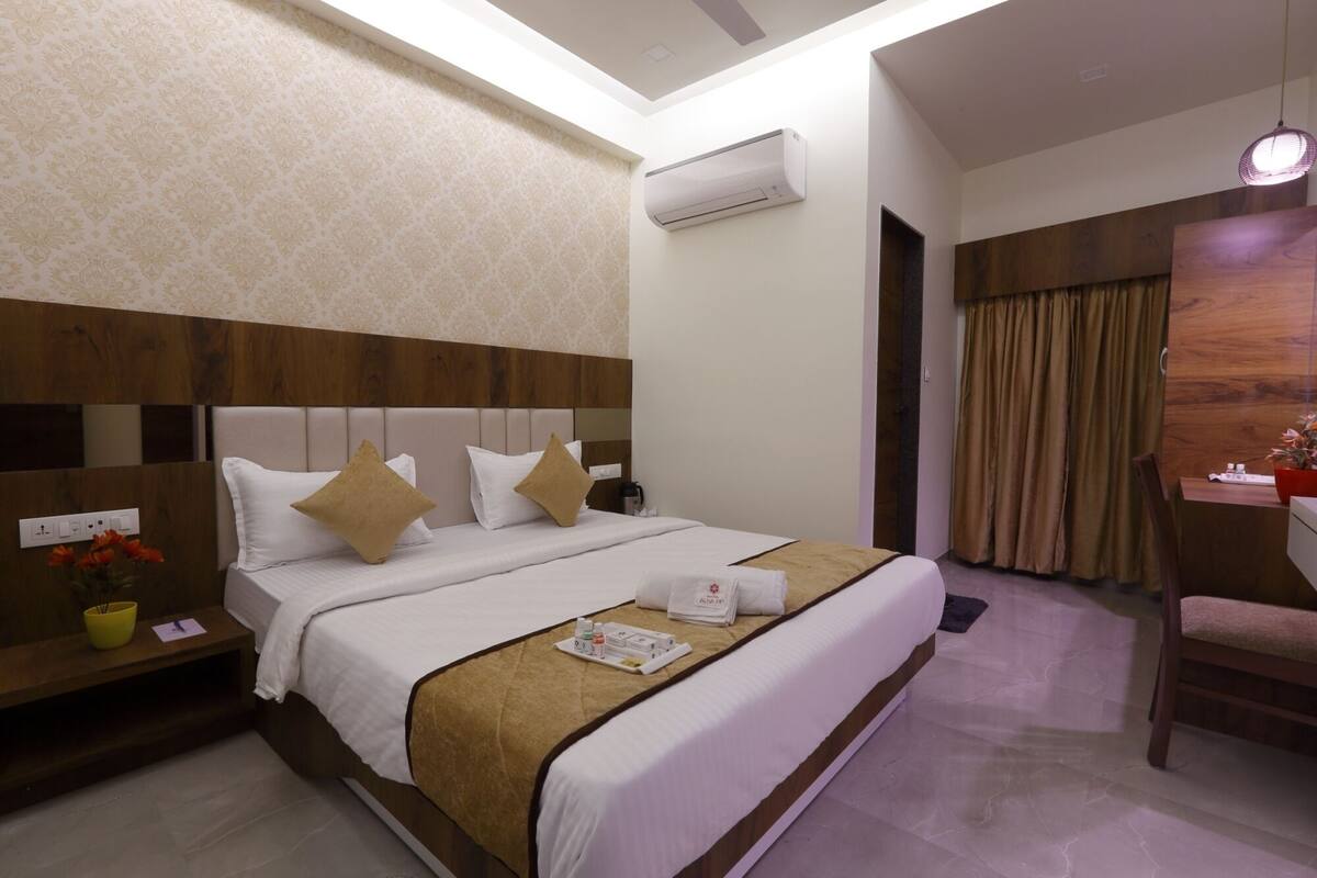 A hotel room with a large bed, a wooden headboard, an air conditioning unit, a desk with a chair, and a window with curtains.