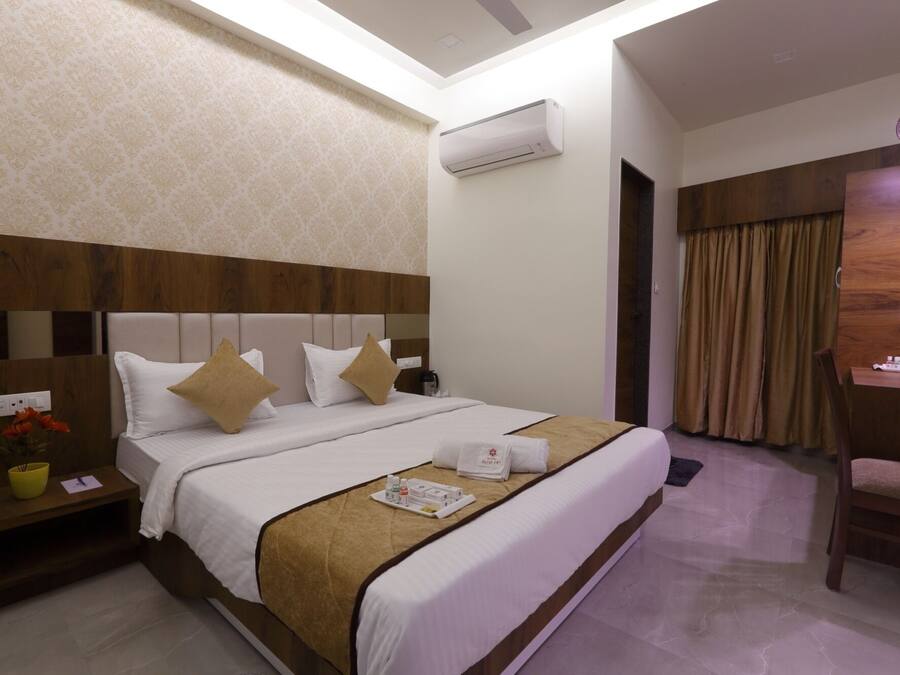 A hotel room with a large bed, a wooden headboard, an air conditioning unit, a desk with a chair, and a window with curtains.