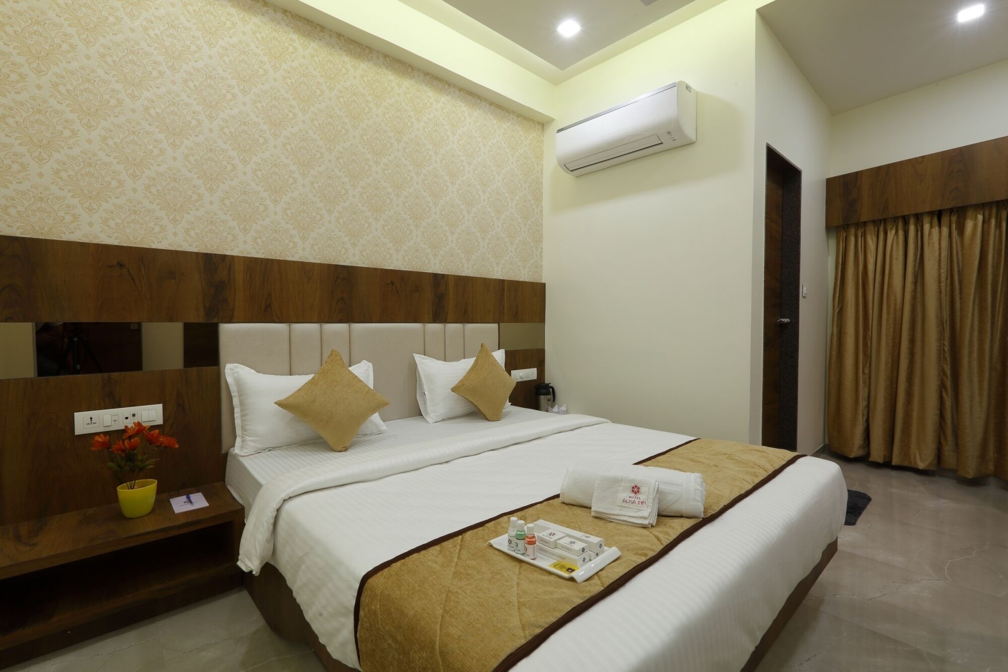 A hotel room with a large bed, a wooden headboard, a small table with a vase of flowers, and an air conditioning unit on the wall.