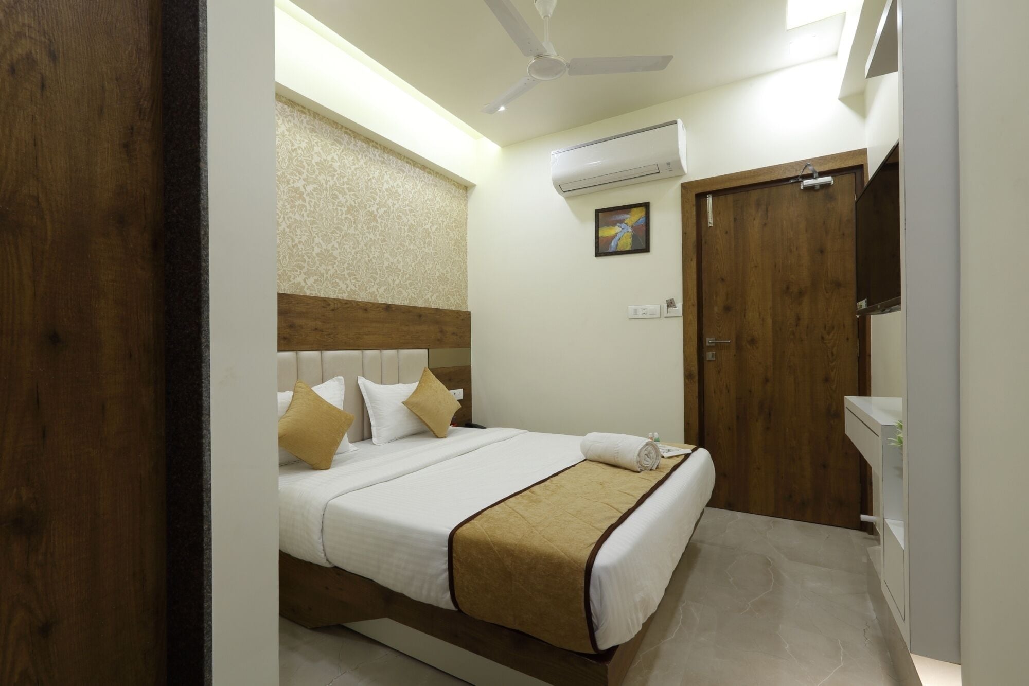 A hotel room with a bed, a ceiling fan, an air conditioning unit, a wooden door, and a small bathroom area.