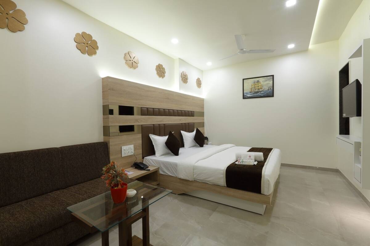 A modern hotel room with a wooden headboard, a bed with white linens, a glass coffee table with a red vase, and a painting on the wall.