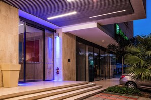 Une entrée moderne avec une porte en verre, des marches menant à un espace d’accueil et une voiture garée à l’extérieur.