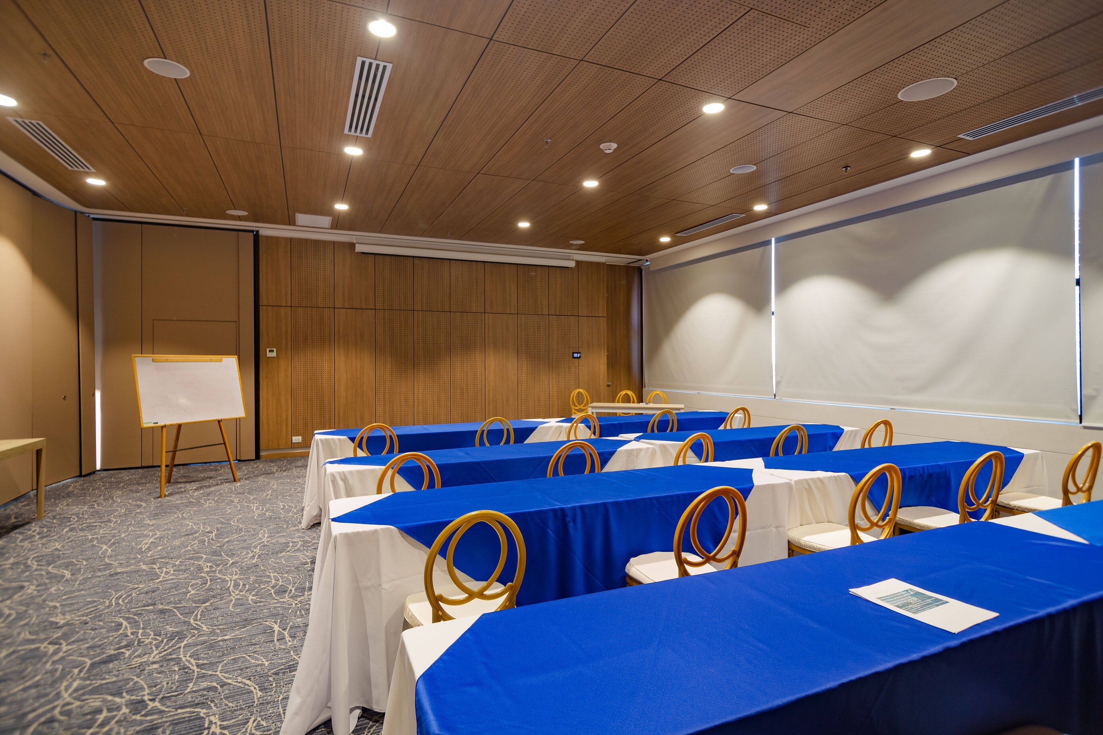 A conference room with tables covered in blue tablecloths, white chairs with gold headrests, and a whiteboard on a stand.