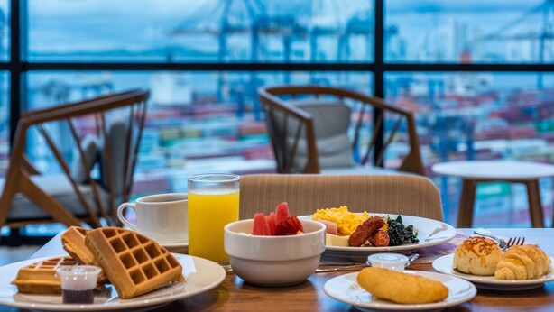 Ein Esstisch mit Waffeln, Obst, Eiern und Croissants, mit Blick auf einen Hafen und im Hintergrund darunter befindliche Kräne.