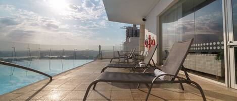A rooftop pool area with lounge chairs and a glass balustrade overlooking a cityscape.