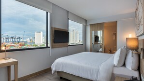 A hotel room with a large bed, a desk with a lamp, a TV mounted on the wall, and a view of the cityscape and port through the window. - Holiday Inn Express Cartagena Manga by IHG (Cartagena)