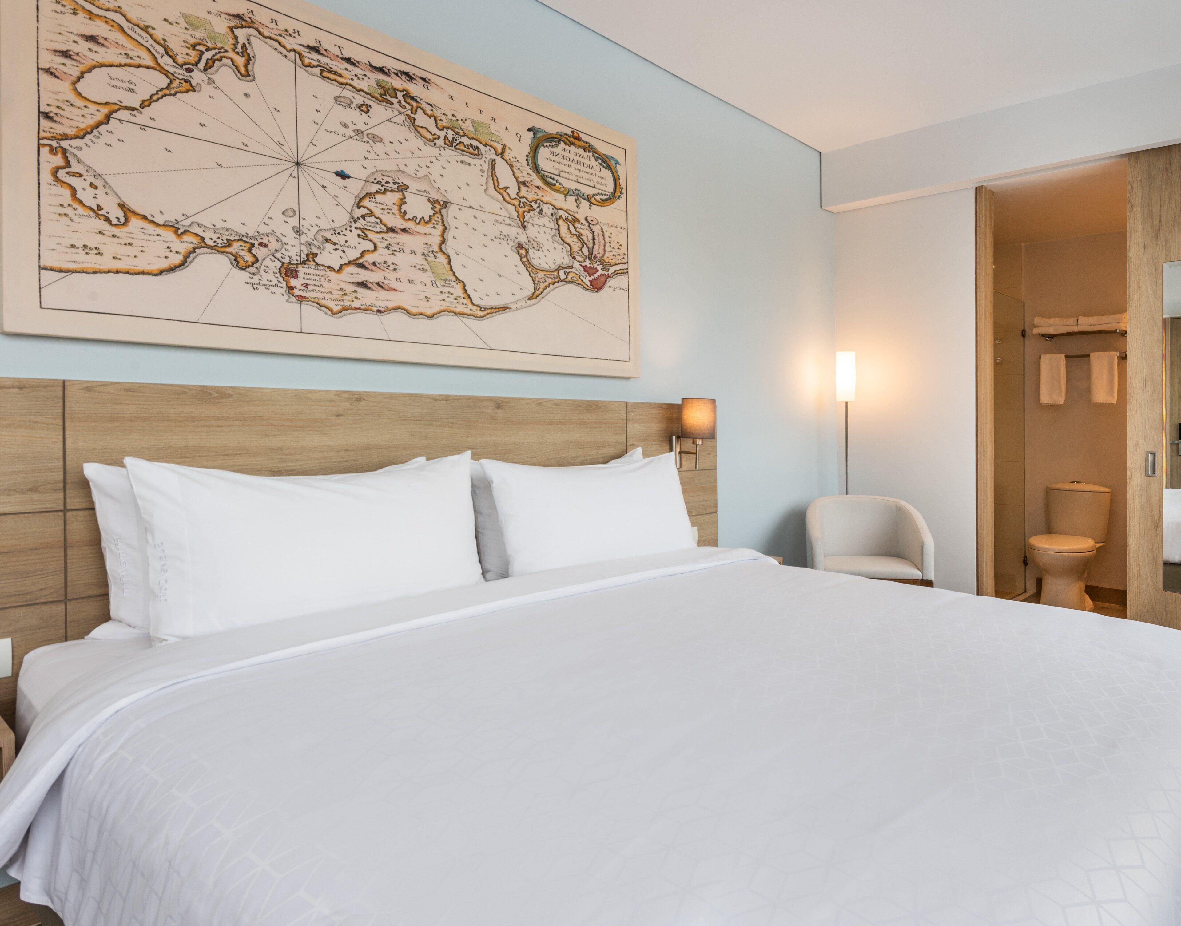 Une chambre d’hôtel avec un grand lit, une tête de lit en bois, une carte accrochée au mur, une chaise et une salle de bain visible à travers une porte ouverte.
