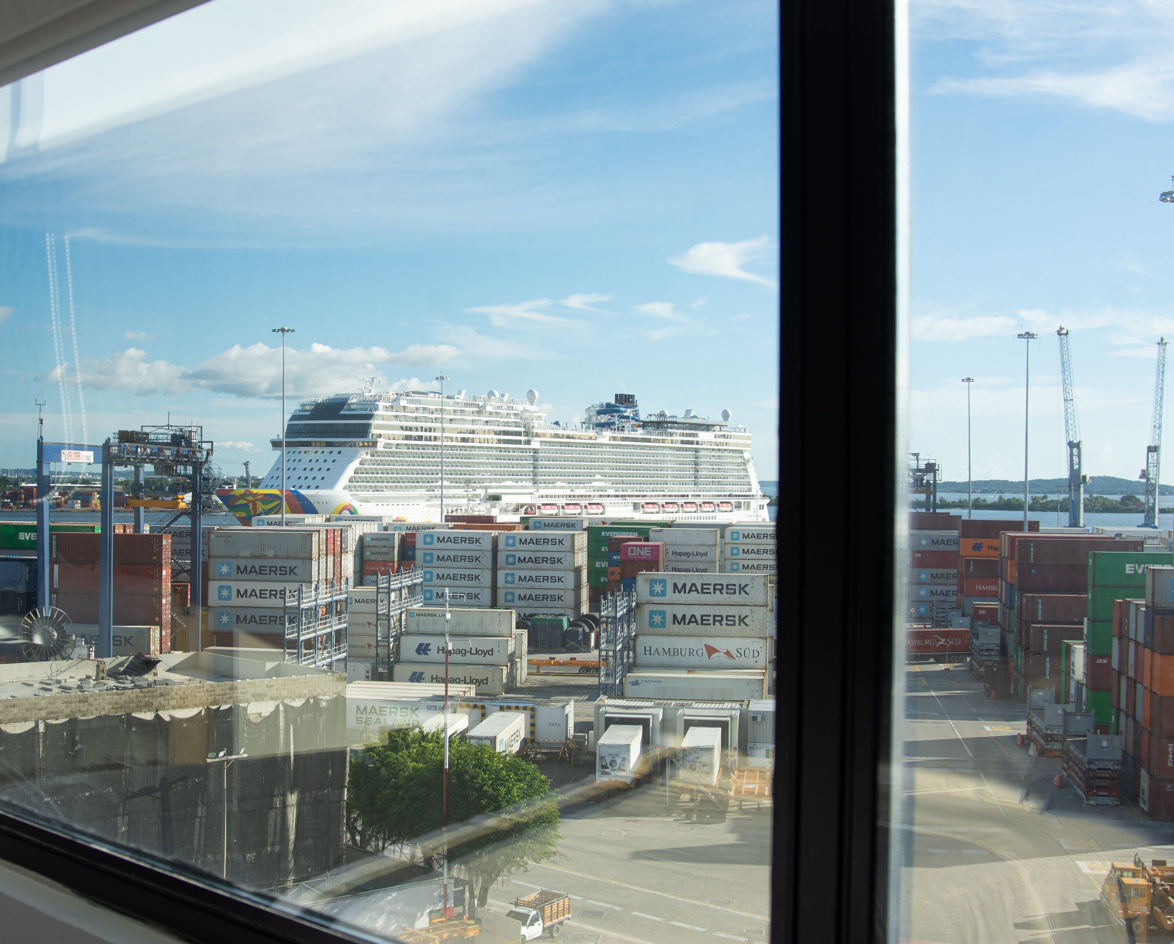 Blick auf ein Kreuzfahrtschiff, das an einem Hafen vor Anker liegt, mit Containern und Kränen im Hintergrund.