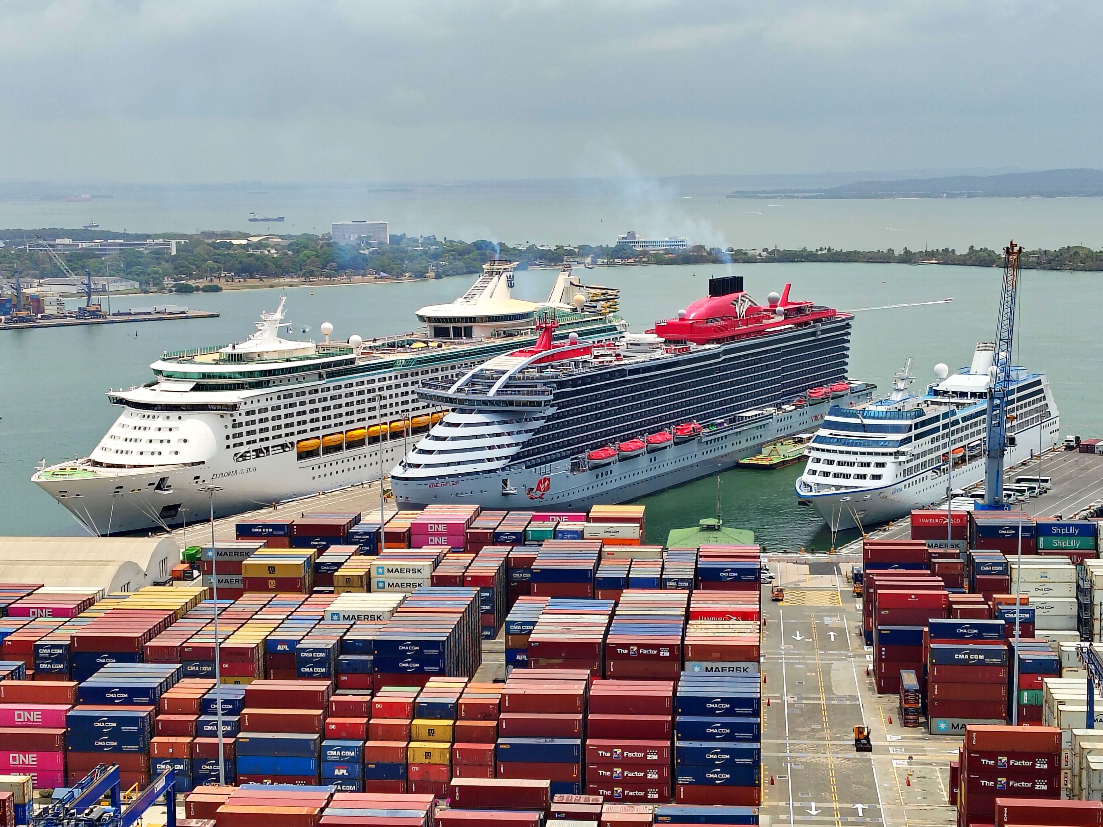 A port with multiple cruise ships docked and surrounded by stacked shipping containers.