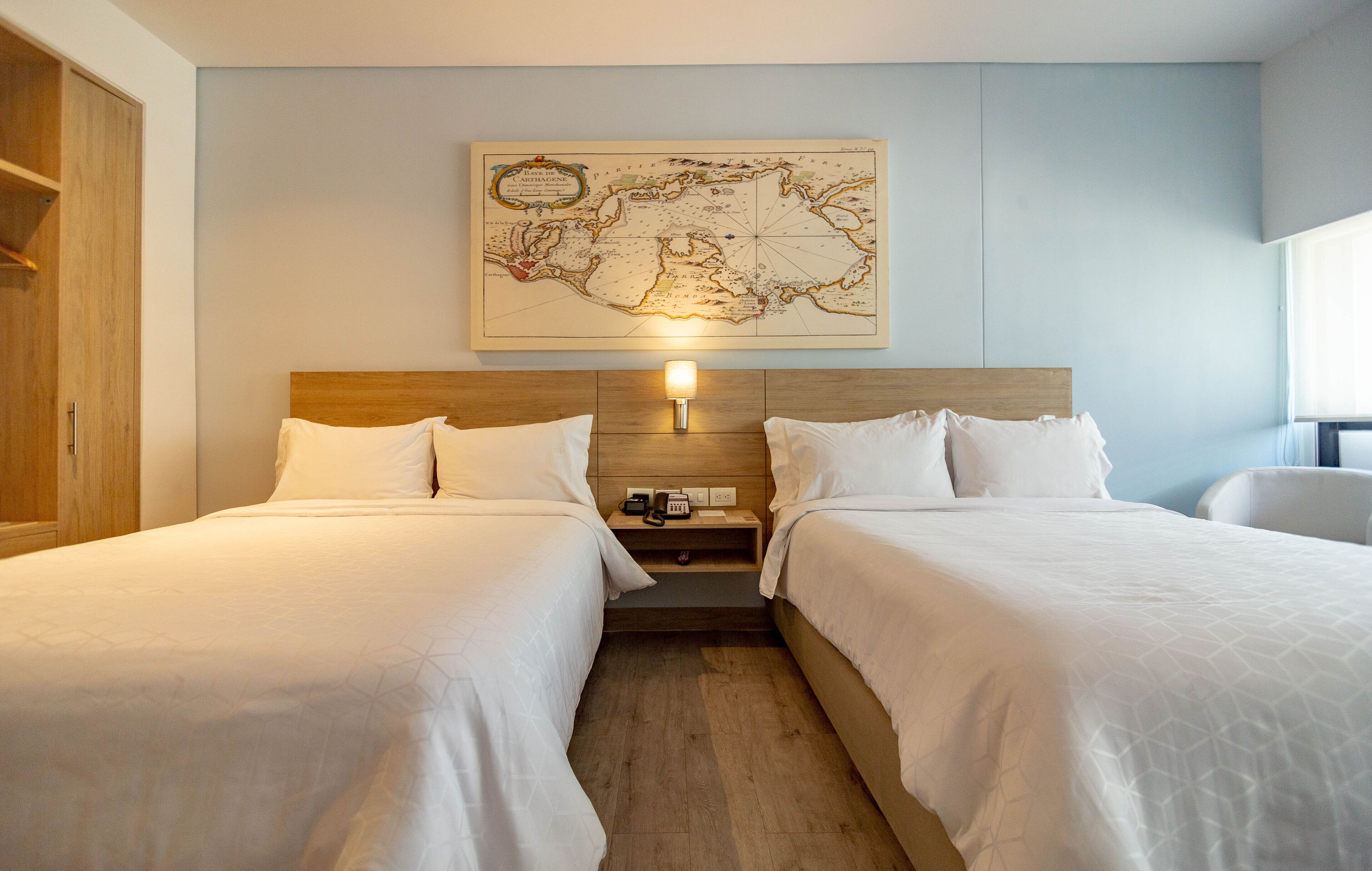 Une chambre d’hôtel avec deux lits, une tête de lit en bois, une carte accrochée au mur et une table de chevet avec une lampe.