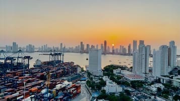 Un puerto con contenedores de carga y grúas, una silueta urbana con rascacielos y una puesta de sol al fondo.