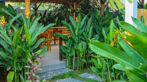 A garden pathway leading to a thatched-roof structure with wooden furniture inside. - Cozy Cottages Lombok (Senggigi)