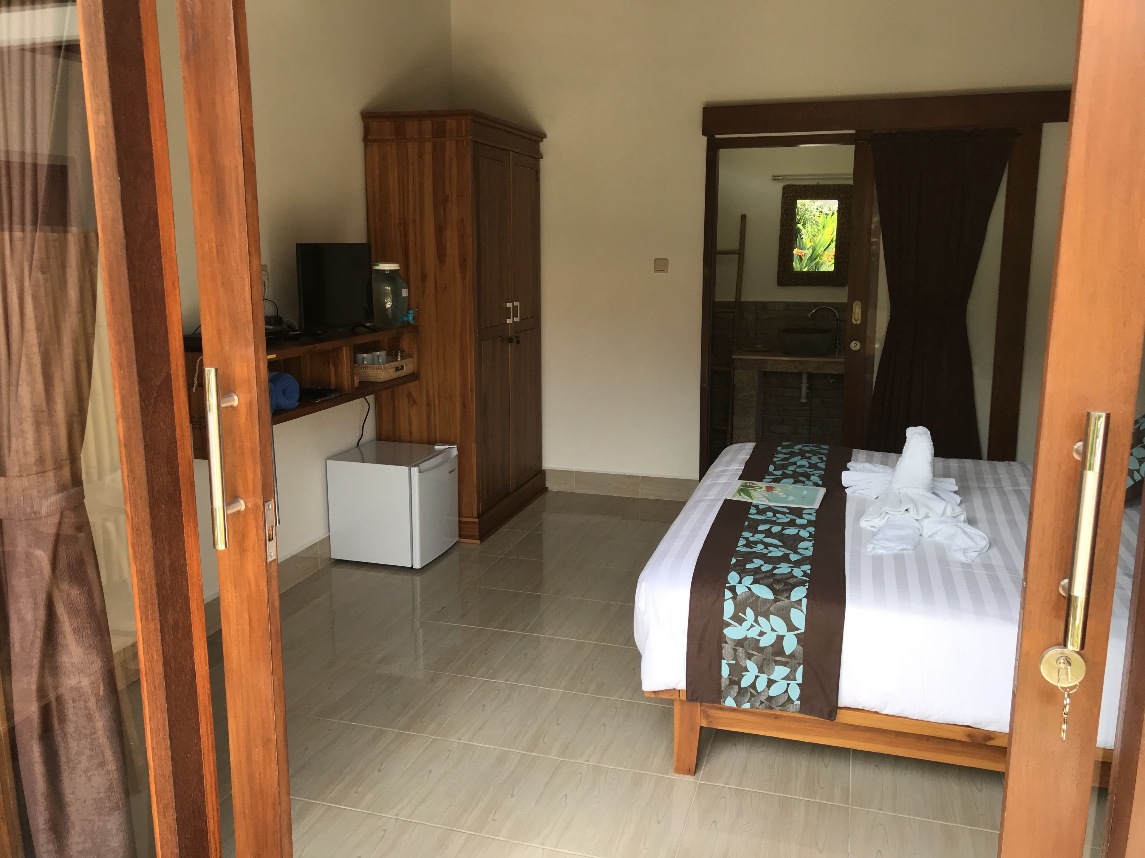 Habitación de hotel con cama, armario de madera, refrigerador blanco pequeño, televisión y baño a la vista a través de una puerta abierta.