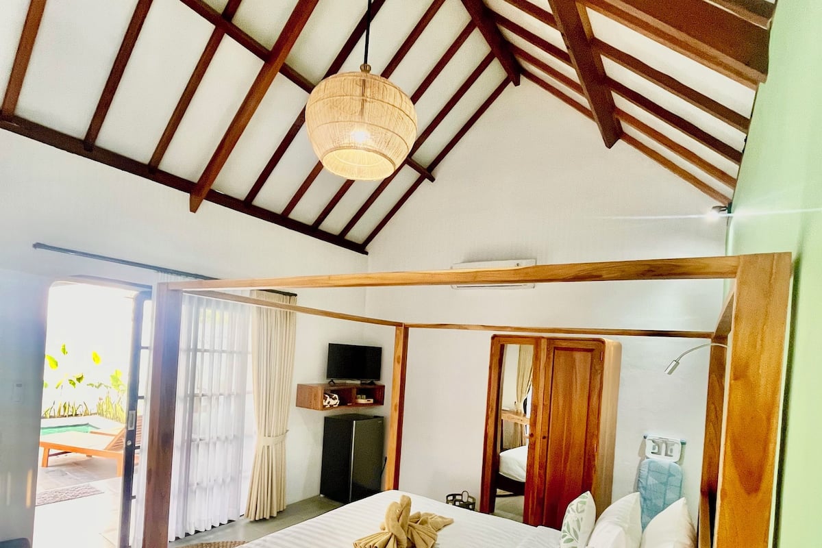 A spacious bedroom with a large bed, wooden beams, a ceiling fan, and a view of the outdoors.