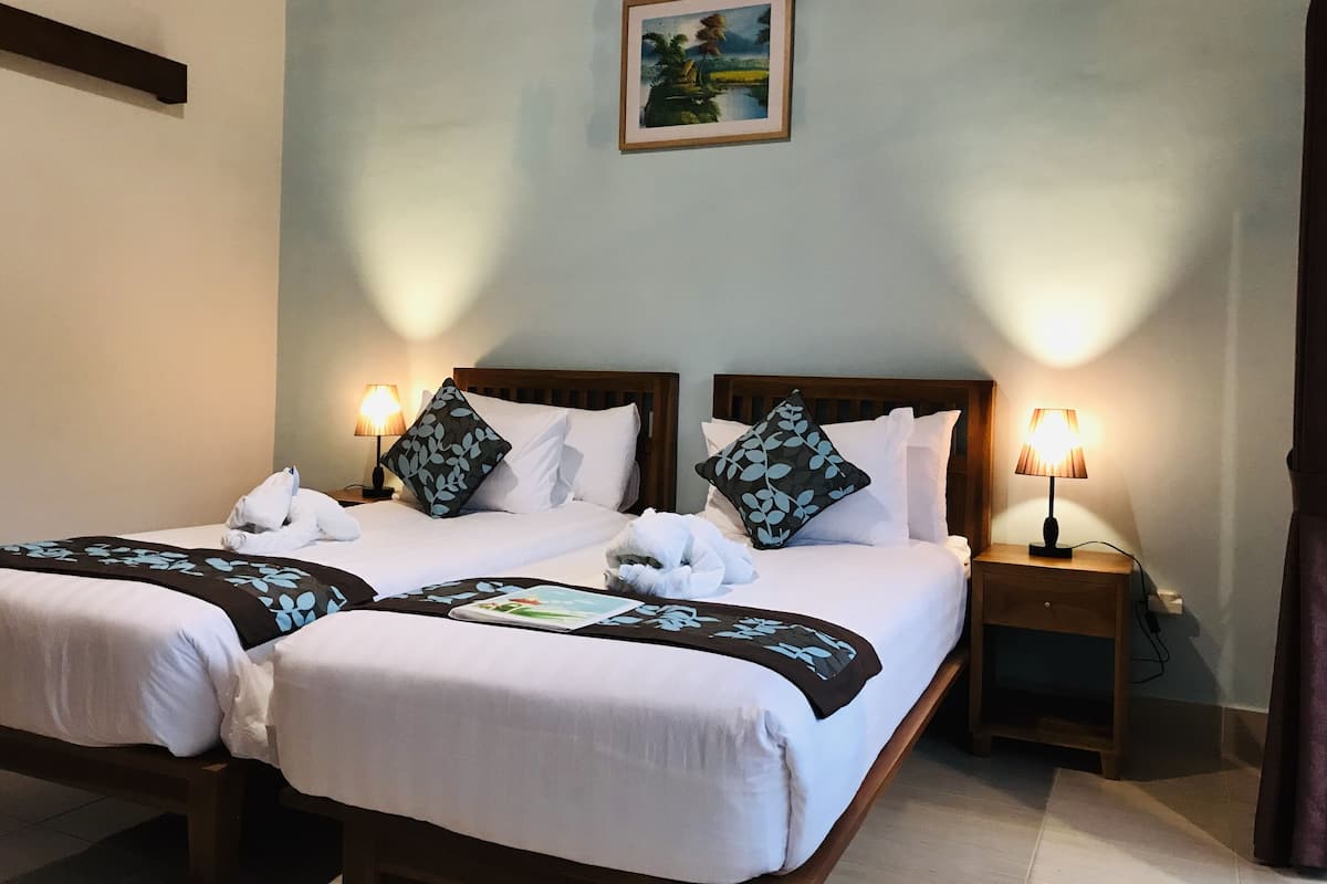 A hotel room with two beds, each with white linens and decorative pillows. There are bedside tables with lamps, a painting on the wall, and a window with curtains.