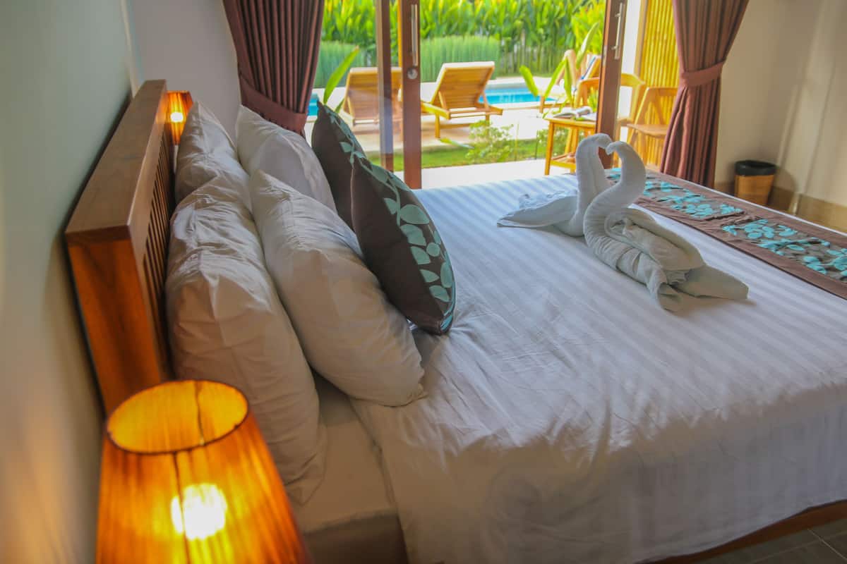 A neatly made bed with pillows and a swan-shaped towel, a lamp, and a view of a pool and chairs outside.