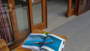 A wooden table with a magazine and a green leaf on it, a bed with white bedding and a blue pillow, and a glass door leading to a room with a bed and a lamp. - Cozy Cottages Lombok (Senggigi)